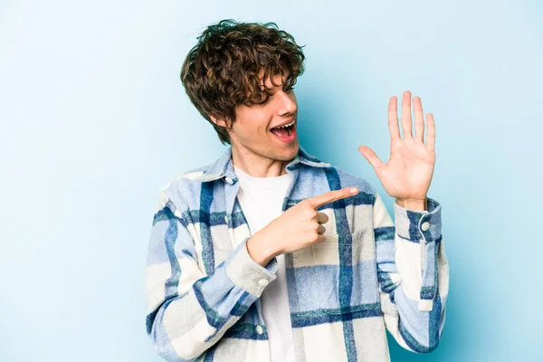 Mavi arka planda izole edilmiş genç beyaz adam gülümseyerek beş numarayı parmaklarıyla gösteriyor..