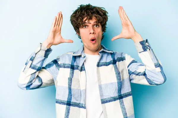Mavi arka planda izole edilmiş genç beyaz bir adam zaferi ya da başarıyı kutluyor, şaşırıyor ve şok oluyor..