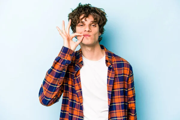 Genç beyaz adam mavi arka planda izole edilmiş ve parmakları dudaklarında bir sır saklıyor..