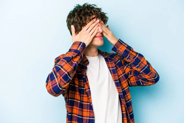 Mavi arka planda izole edilmiş genç beyaz adam elleriyle gözlerini kapatmaktan korkuyor..