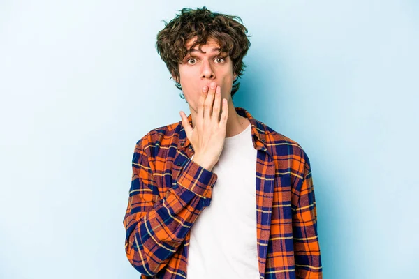 Mavi arka planda izole edilmiş genç beyaz adam şok olmuş, ağzı ellerle kaplanmış, yeni bir şey keşfetmeye hevesli..