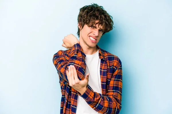 Kafkasyalı genç adam mavi arka planda izole edilmiş dirsek masajı yapıyor, kötü bir hareketin ardından acı çekiyor..