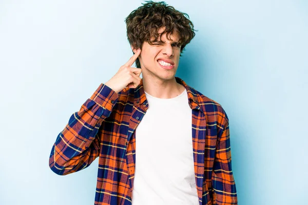 Genç beyaz adam mavi arka planda izole edilmiş parmaklarıyla kulaklarını kapatıyor, gürültülü bir ortam tarafından stresli ve umutsuz..