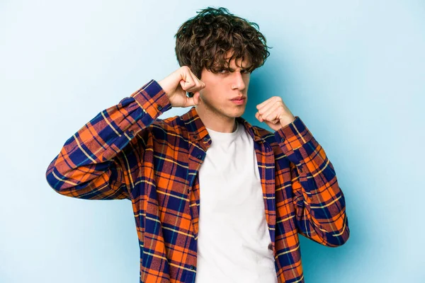 Kafkasyalı genç bir adam mavi arka planda izole edilmiş. Yumruk atıyor, sinirleniyor, kavga ediyor, kavga ediyor..