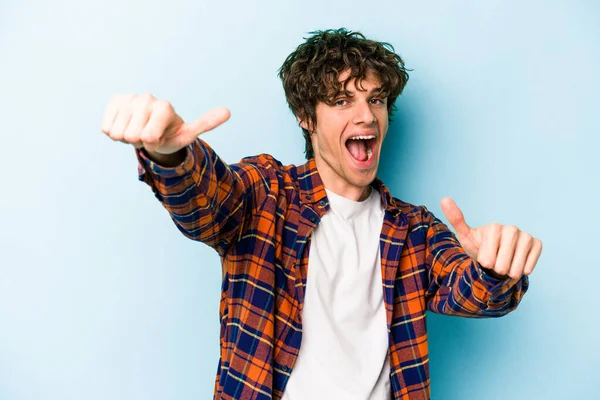 Kafkasyalı genç adam mavi arka planda izole edilmiş. Baş parmağını kaldırıyor, gülümsüyor ve kendinden emin..