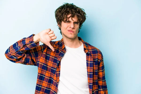 Mavi arka planda izole edilmiş genç beyaz bir adam baş parmağını aşağı gösteriyor ve hoşlanmadığını ifade ediyor..