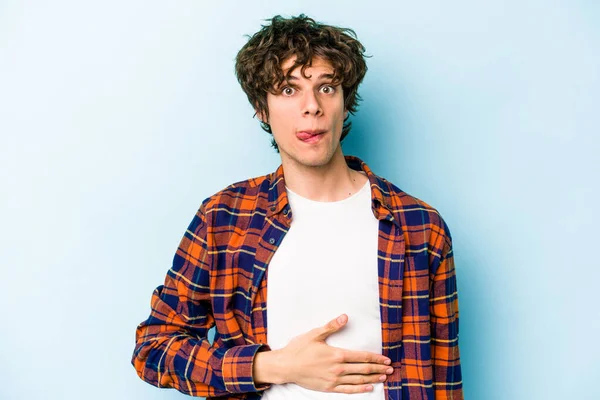 Mavi arka planda izole edilmiş genç beyaz adam karnına dokunuyor, nazikçe gülümsüyor, yemek yiyor ve tatmin oluyor..