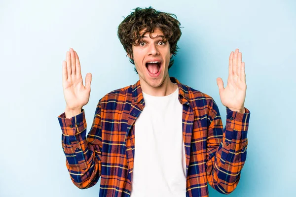 Mavi arka planda izole edilmiş genç beyaz adam hoş bir sürpriz alıyor, heyecanlı ve el kaldırıyor..