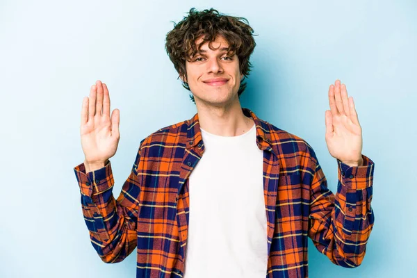 Kafkasyalı genç adam mavi arka planda izole edilmiş. Elinde işaret parmaklarıyla küçük bir şey tutuyor, gülümsüyor ve kendinden emin..