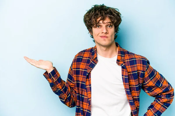 Mavi arka planda izole edilmiş genç beyaz bir adam bir palmiye üzerinde fotokopi alanı ve belinde bir el daha gösteriyor..