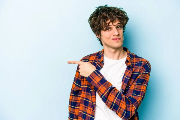 Mavi arka planda izole edilmiş genç beyaz adam gülümsüyor ve boş alanda bir şeyler gösteriyor..