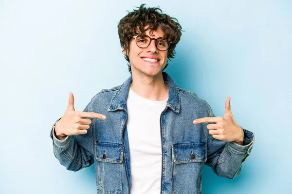 Mavi arka planda izole edilmiş genç beyaz adam bir gömlek fotokopi alanını işaret ediyor, gururlu ve kendinden emin.