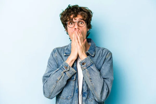 Mavi arka planda izole edilmiş genç beyaz adam elleriyle ağzını kapatırken şok oldu..