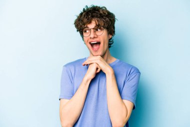 Kafkasyalı genç adam mavi arka planda izole edilmiş şans için dua ediyor, şaşırmış ve ağzını açıp önüne bakıyor..