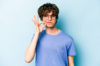 Genç beyaz adam mavi arka planda izole edilmiş ve parmakları dudaklarında bir sır saklıyor..