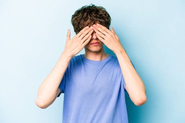 Mavi arka planda izole edilmiş genç beyaz adam elleriyle gözlerini kapatmaktan korkuyor..