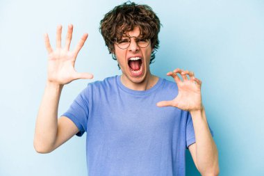 Mavi arka planda izole edilmiş genç beyaz adam pençelerini kedi taklidi yaparken gösteriyor, agresif bir hareket..