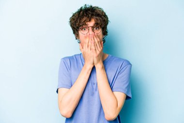 Mavi arka planda izole edilmiş genç beyaz adam şok olmuş, ağzı ellerle kaplanmış, yeni bir şey keşfetmeye hevesli..