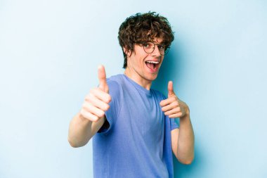Kafkasyalı genç adam mavi arka planda izole edilmiş. Baş parmağını kaldırıyor, gülümsüyor ve kendinden emin..