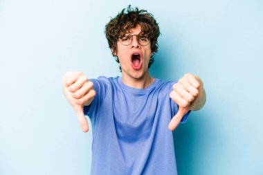Mavi arka planda izole edilmiş genç beyaz bir adam baş parmağını aşağı gösteriyor ve hoşlanmadığını ifade ediyor..