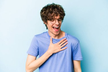 Mavi arka planda izole olmuş genç beyaz adam yüksek sesle gülüyor ve elini göğsüne koyuyor..
