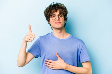 Mavi arka planda izole edilmiş genç beyaz adam karnına dokunuyor, nazikçe gülümsüyor, yemek yiyor ve tatmin oluyor..