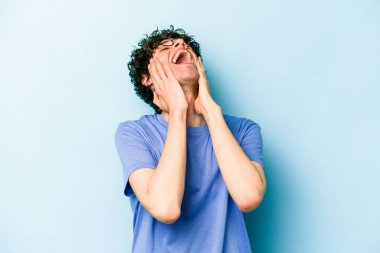 Mavi arka planda izole edilmiş genç beyaz adam ellerini başının üstünde tutarak neşe içinde gülüyor. Mutluluk kavramı.