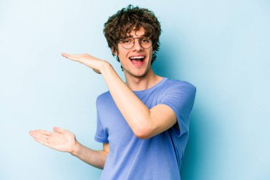 Kafkasyalı genç adam mavi arka planda izole edilmiş. Elinde işaret parmaklarıyla küçük bir şey tutuyor, gülümsüyor ve kendinden emin..