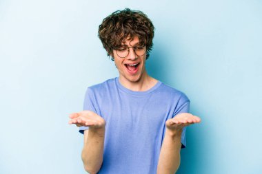 Kafkasyalı genç adam mavi arka planda izole edilmiş. Elinde avuç içi olan bir şey var ve kameraya sunuyor..