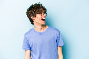 Kafkasyalı genç adam mavi arka planda izole edilmiş rahat ve mutlu bir şekilde gülüyor, boynunda dişlerini gösteriyor..