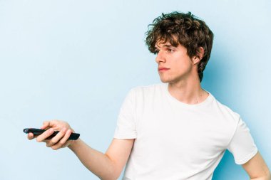 Elinde televizyon kumandası olan beyaz bir adam mavi arka planda izole edilmiş.