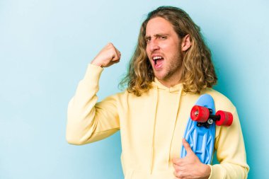 Genç kaykaycı beyaz adam mavi arka planda izole edilmiş zafer kazandıktan sonra yumruğunu kaldırıyor, kazanan konsept.