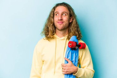 Genç kaykaycı beyaz adam mavi arka planda izole edilmiş hedeflerine ve amaçlarına ulaşma hayali kuruyor.