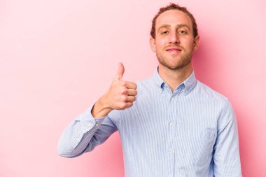 Pembe arka planda izole edilmiş genç beyaz adam gülümsüyor ve başparmağını kaldırıyor.