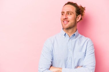 Pembe arka planda izole edilmiş genç beyaz adam kollarını kavuşturarak gülümsüyor..