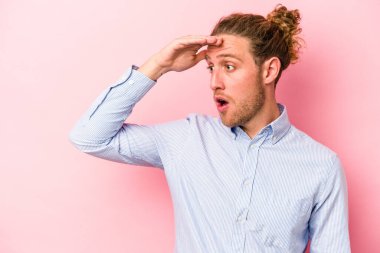 Pembe arka planda izole edilmiş genç beyaz adam uzaklara bakıyor ve elini alnına koyuyor..