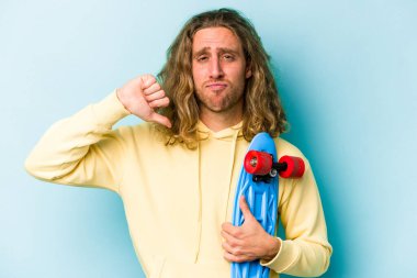 Genç kaykaycı beyaz adam mavi arka planda izole edilmiş, hoşlanmadığı bir hareket gösteriyor, başparmakları aşağıda. Anlaşmazlık konsepti.