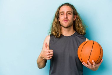 Basketbol oynayan beyaz bir adam mavi arka planda izole edilmiş gülümsüyor ve başparmağını kaldırıyor.