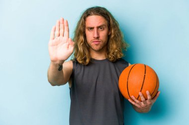 Mavi arka planda izole bir şekilde basketbol oynayan genç beyaz adam elini uzatmış, dur işaretini göstererek seni engelliyor..