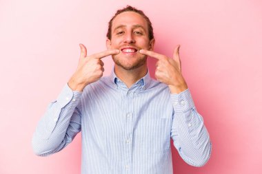 Pembe arka planda izole edilmiş genç beyaz adam gülümsüyor, parmaklarını ağıza doğrultuyor..