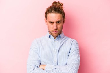 Pembe arka planda izole edilmiş genç beyaz adam hoşnutsuzca kaşlarını çatmış, kollarını kavuşturmuş..