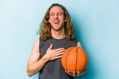 Mavi arka planda basketbol oynayan beyaz bir genç elini göğsünde tutarak kahkahalar atıyor..