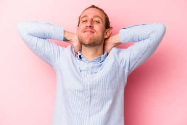 Pembe arka planda izole edilmiş genç beyaz bir adam kendini güvende hissediyor, elleri başının arkasında..