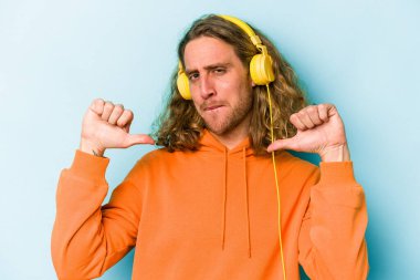 Mavi arka planda izole edilmiş müzik dinleyen genç beyaz adam gururlu ve kendinden emin hissediyor, örnek olarak..