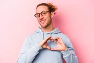 Pembe arka planda izole edilmiş genç beyaz adam gülümsüyor ve elleriyle kalp şeklini gösteriyor..