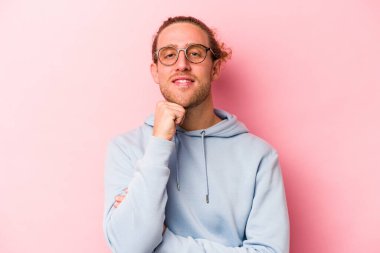 Pembe arka planda izole edilmiş genç beyaz adam gülümsüyor ve kendinden emin, elleriyle çenesine dokunuyor..