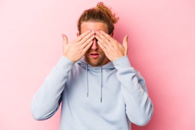 Pembe arka planda izole edilmiş genç beyaz adam elleriyle gözlerini kapatmaktan korkuyor..