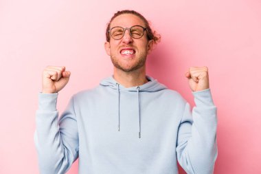 Pembe arka planda izole edilmiş genç beyaz bir adam zaferi, tutkuyu ve coşkuyu kutluyor, mutlu bir ifade..