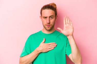 Pembe arka planda izole edilmiş genç beyaz adam yemin ediyor, elini göğsüne koyuyor..