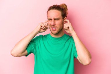 Pembe arka planda izole edilmiş genç beyaz bir adam parmaklarıyla kulaklarını kapatıyor, gürültülü bir ortam yüzünden stresli ve çaresiz..